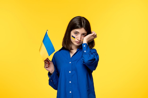 Ukraine russian conflict young pretty girl flags on cheeks yellow background wiping tears off