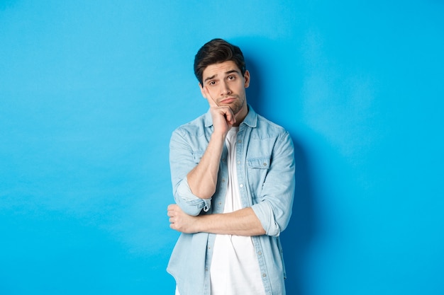 Unamused and bored man looking without interest at camera, standing against bllue background.