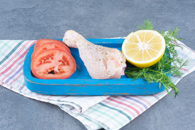 Uncooked chicken leg, tomato and lemon slice on blue plate.