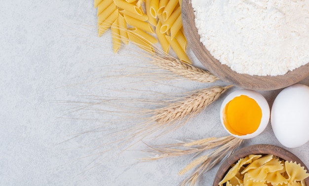 Uncooked different types of pasta with flour and raw egg on marble surface.