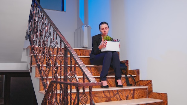 Unemployed businesswoman holding box of stuff on company stairs after beeing fired from the job. Colleagues leaving office building. Unemployed depressed woman lost her workplace