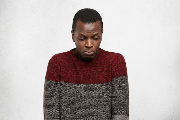 Unhappy Afro American student feeling uncomfortable and ashamed, looking down with sad expression while having troubles at college. Portrait of sad young black male with no motivation and enegry