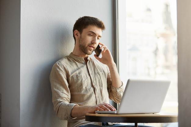 Unhappy handsome guy with dark hair, sitting in cafe, working on laptop computer and talking with dissatisfied customer on phone.