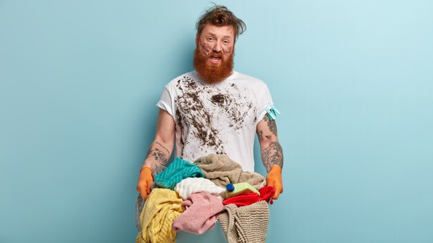 Unhappy indignant redhead man with thick bristle, wears untidy t shirt, rubber gloves, carries heap of laundry, has dirty face, stands against blue wall, isnt eager to do washing at home