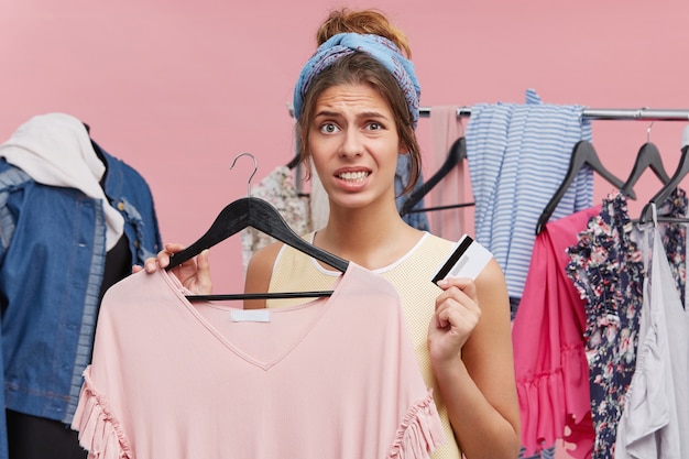 Unhappy woman doing shopping, standing in clothes shop, holding new dress and credit card, being shot of money, having financial crisis, wanting to buy new clothes immediately. Shopping expanses