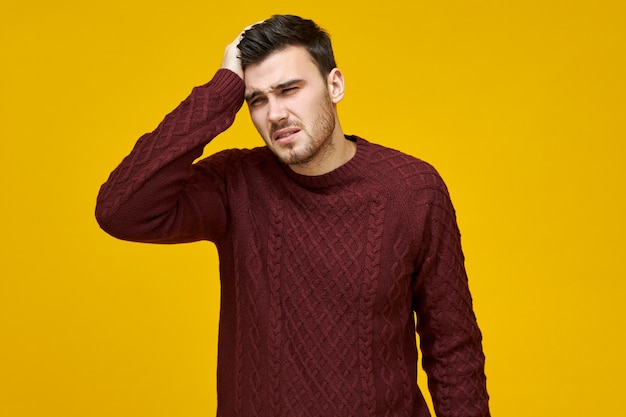  unhappy young man in warm sweater having cold, flu or bad day at work suffering splitting headache, holding hand son his head, needs anti febrile medicine to get down temperature