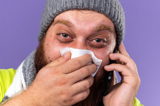 Unhealthy bearded man in hat and with warm scarf around neck feeling terrible suffering from flu talking on mobile phone blowing running nose sneezing in tissue standing over purple wall