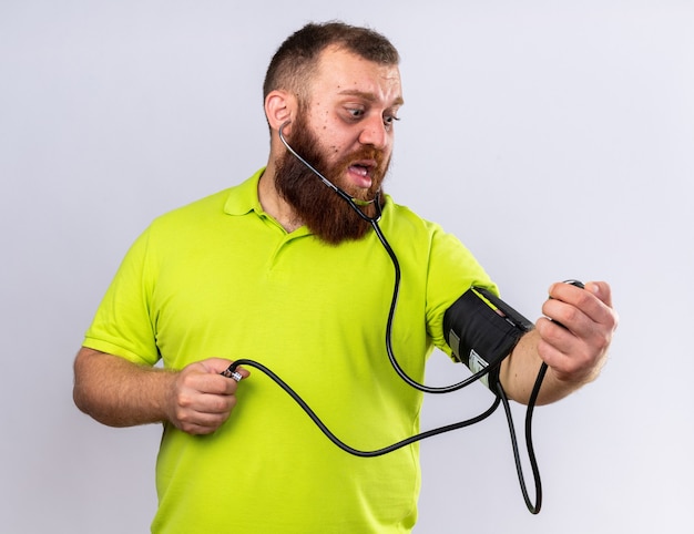 Unhealthy bearded man in yellow polo shirt feeling sick measuring blood pressure using tonometer looking worried standing over white wall