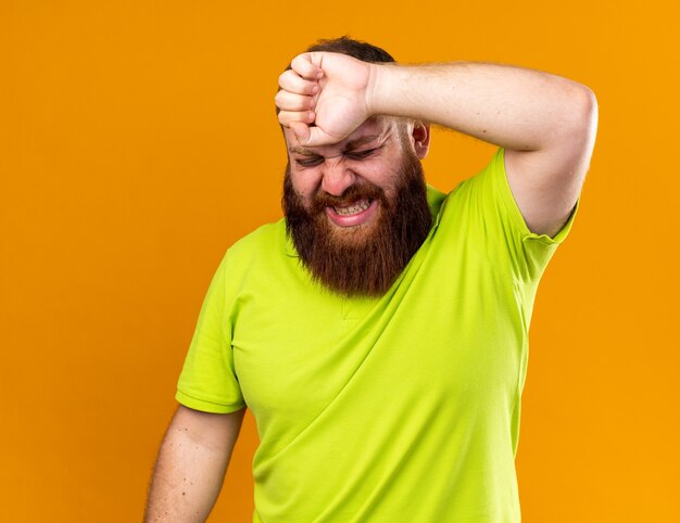 Unhealthy bearded man in yellow polo shirt feeling terrible suffering from cold touching his forehead suffering from strong headache feeling pain