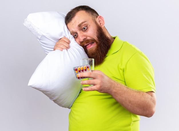 Unhealthy bearded man in yellow polo shirt with pillow holding glass of water and pills feeling sick suffering from flu standing over white wall