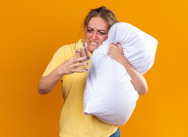 Unhealthy woman in yellow shirt suffering from flu and cold feeling unwell hugging pillow holding pills and glass of water looking annoyed and disappointed standing over orange wall