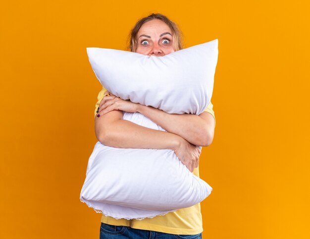 Unhealthy woman in yellow shirt suffering from flu and cold feeling unwell hugging pillow  scared standing over orange wall