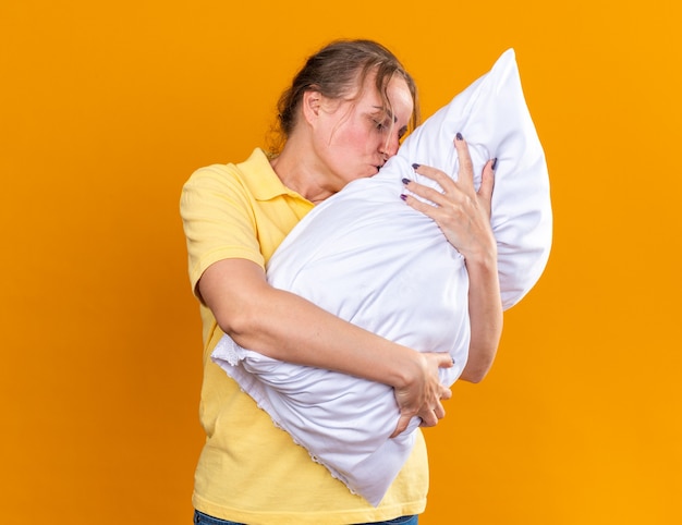 Unhealthy woman in yellow shirt suffering from flu and cold hugging pillow feeling better kissing pillow standing over orange wall