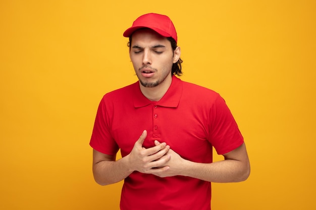 unhealthy young delivery man wearing uniform and cap putting hands on chest while keeping eyes closed isolated on yellow background