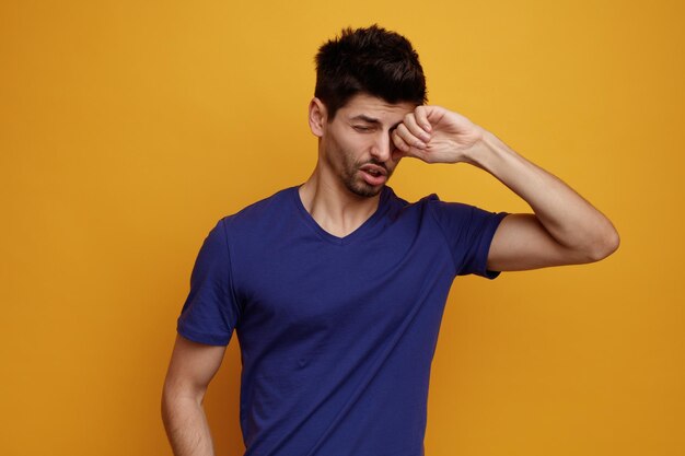 Unhealthy young handsome man keeping hand on eye with closed eyes on yellow background