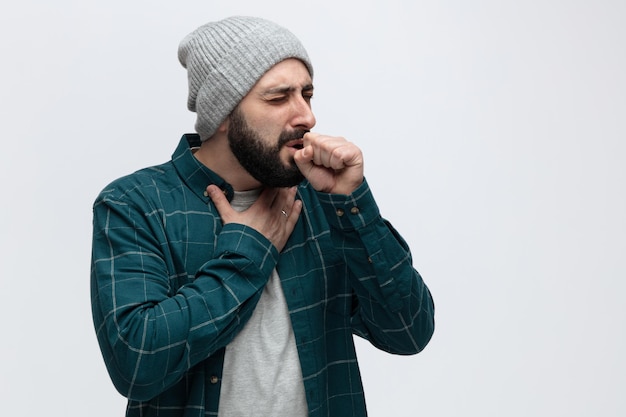 Free photo unhealthy young man wearing winter hat keeping hand on chest and fist in front of mouth coughing with closed eyes isolated on white background with copy space