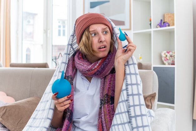 Unhealthy young woman in hat wrapped in blanket feeling unwell and sick holding enemas looking confused having doubts sitting on couch in light living room