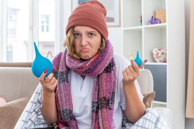 Unhealthy young woman in hat wrapped in blanket feeling unwell and sick holding enemas looking confused having doubts sitting on couch in light living room