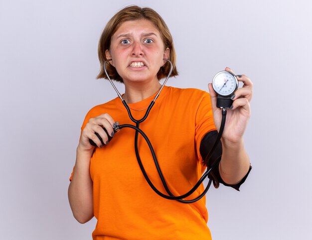 Unhealthy young woman in orange t-shirt feeling unwell measuring blood pressure using tonometer looking worried and disappointed standing over white wall