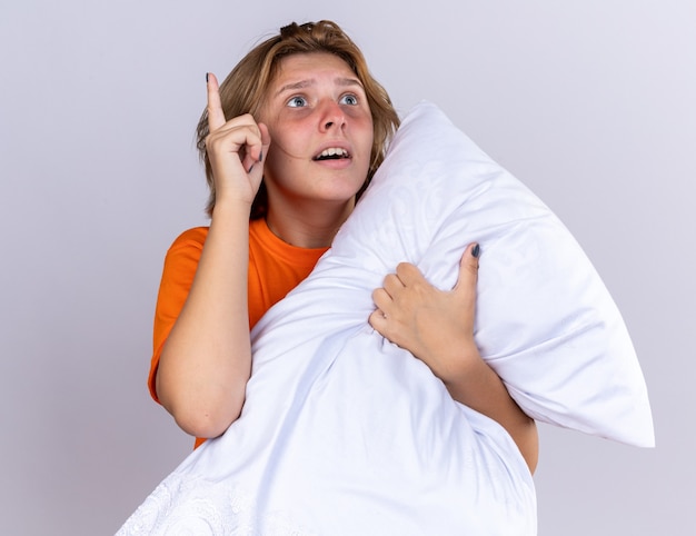 Unhealthy young woman in orange t-shirt holding pillow feeling sick suffering from flu looking up surprised showing index finger having new idea standing over white wall