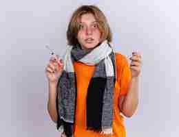 Free photo unhealthy young woman in orange t-shirt with warm scarf feeling terrible suffering from flu holding syringe and ampoule  confused standing over white wall