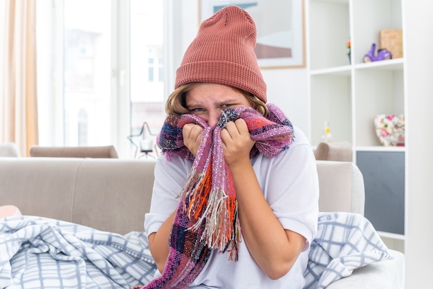 Unhealthy young woman in warm hat with scarf around neck feeling unwell and sick suffering from cold and flu looking worried sitting on couch in light living room