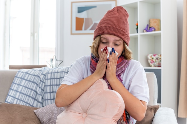 Unhealthy young woman in warm hat with scarf holding pillow blowing nose in tissue suffering from cold and flu sitting on the chair in light living room