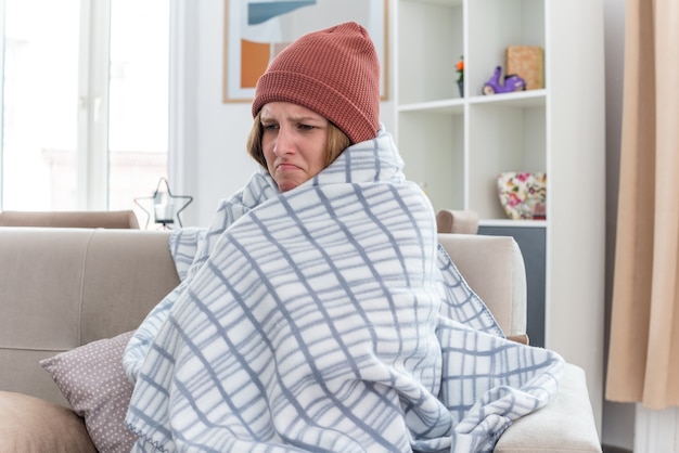 Unhealthy young woman in warm hat wrapped in blanket looking unwell and sick suffering from cold and flu having fever and headache sitting on couch in light living room