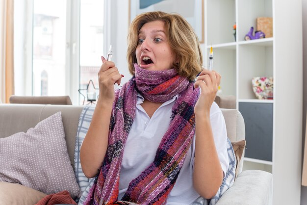 Unhealthy young woman with warm scarf around neck feeling unwell and sick suffering from cold and flu holding syringe and ampoule looking worried and surprised sitting on couch in light living room