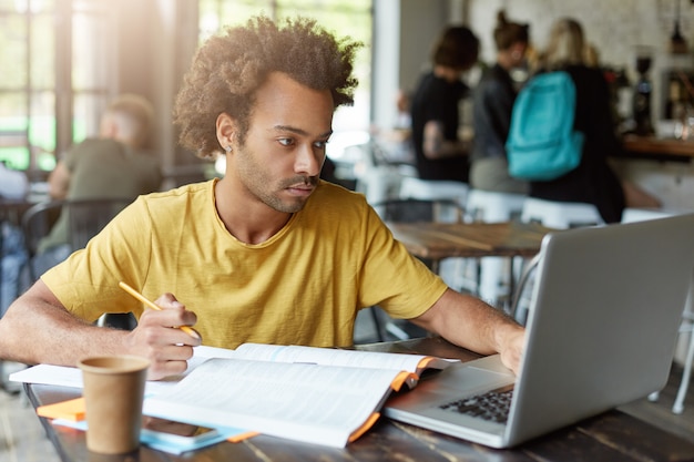 University student with dark skin and African hairstyle sitting at cafe working with books and notebook while getting prepared for exam finding necessary information in internet having serious look