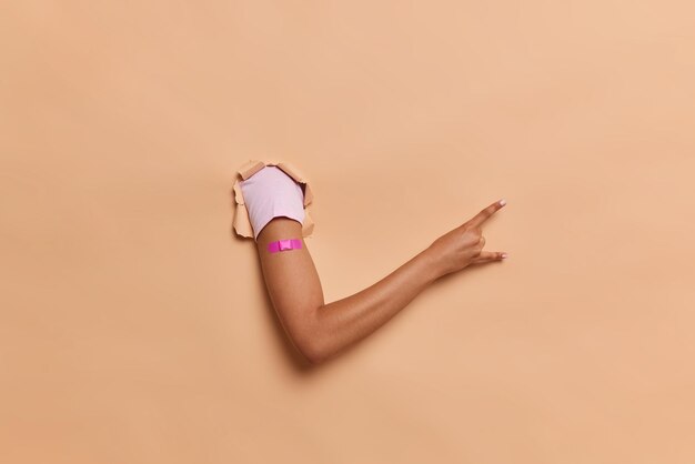 Unknown person shows arm with adhesive plaster after getting vaccination shows rock n roll gesture against brown studio background Vaccine and immunization concept Health care during pandemic