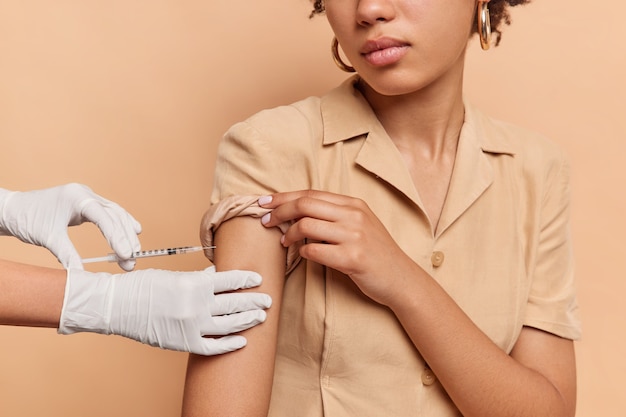 Free Photo unknown person with dark skin makes covid 19 vaccination gets injection dose in shoulder wears brown dress in one tone with wall