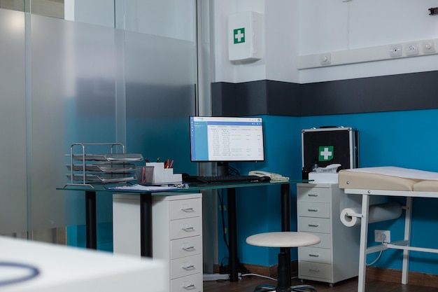 Free photo unoccupied modern clinic workspace with nobody in it having turned on computer screen showing medical report. medical room having firstaid kit, contemporary hospital furniture and patients documents.