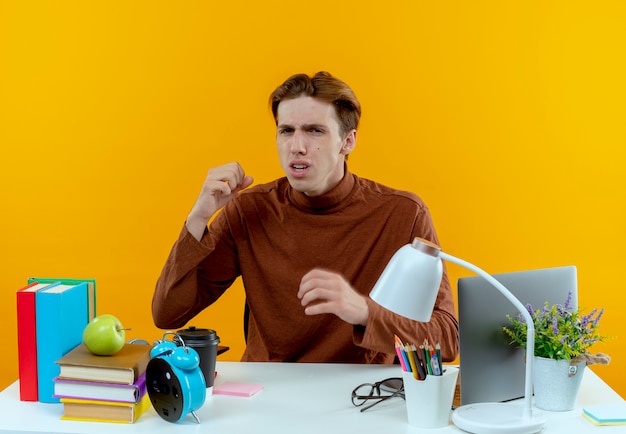 Free photo unpeased young student boy sitting at desk with school tools isolated on yellow wall