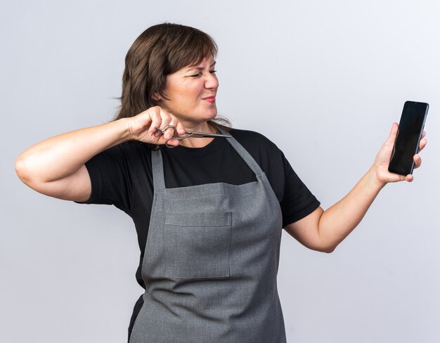 Unpleased adult caucasian female barber in uniform holding scissors and looking at phone isolated on white background with copy space