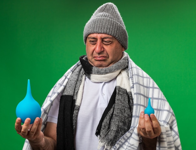 unpleased adult ill caucasian man with scarf around neck wearing winter hat wrapped in plaid holding and looking at enemas isolated on green wall with copy space