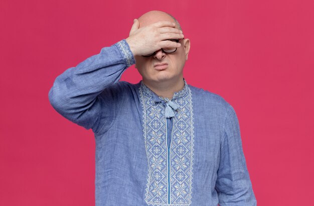 Unpleased adult man in blue shirt wearing glasses putting hand on his forehead 