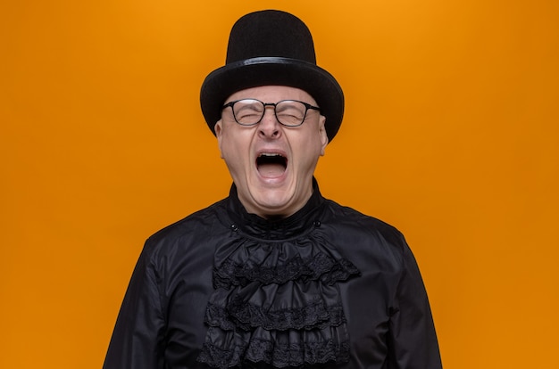 Free photo unpleased adult slavic man with top hat and optical glasses in black gothic shirt screaming standing with closed eyes