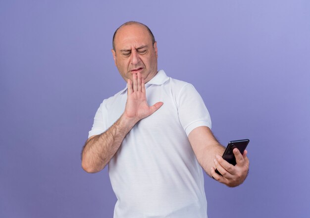 Unpleased casual mature businessman holding and looking at mobile phone and gesturing no with hand isolated on purple background with copy space