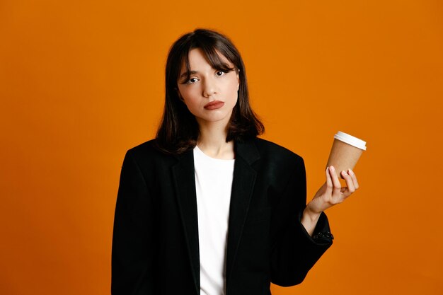 Unpleased holding coffee cup young beautiful female wearing black jacket isolated on orange background