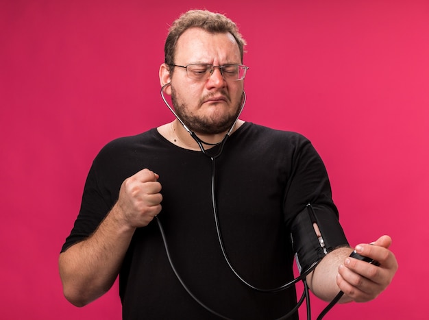 Unpleased middle-aged ill male measuring his own pressure with sphygmomanometer isolated on pink wall