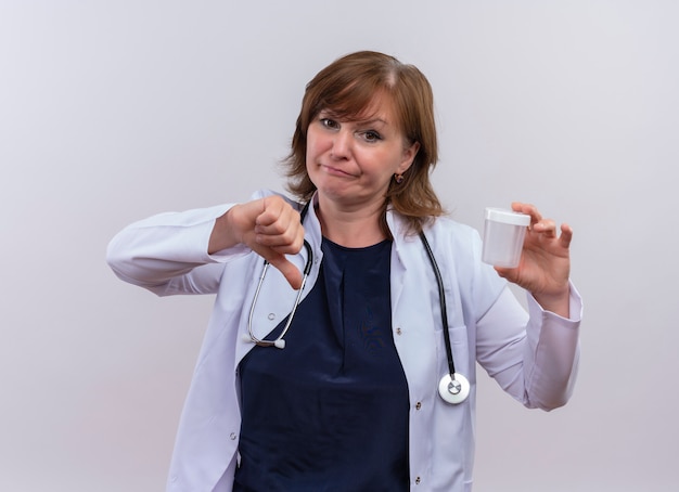 Unpleased middle-aged woman doctor wearing medical robe and stethoscope holding medical beaker and showing thumb down on isolated white background