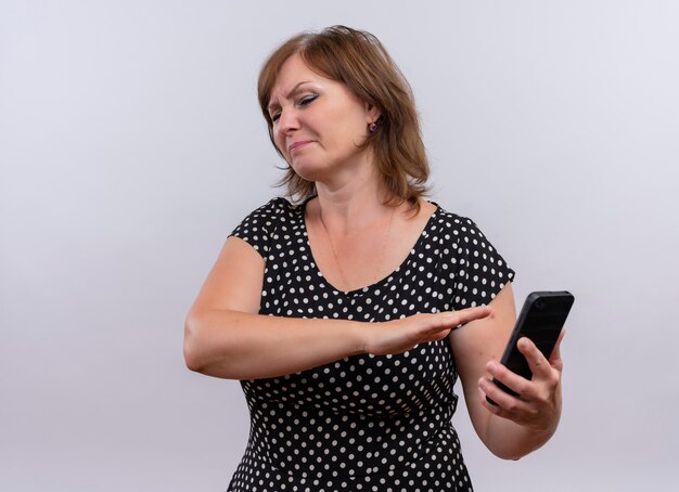 Unpleased middle-aged woman holding mobile phone and pointing with hand at it on isolated white background