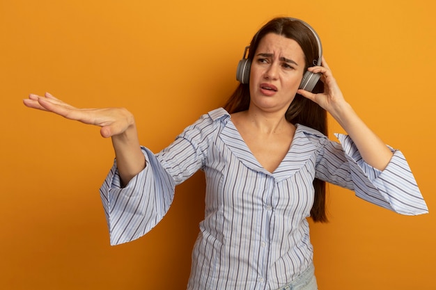 Unpleased pretty caucasian woman on headphones holds hand open and looks at side on orange