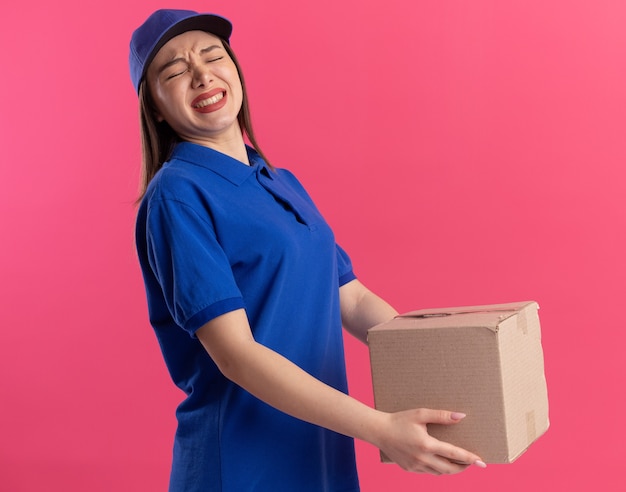 Unpleased pretty delivery woman in uniform holds cardbox isolated on pink wall with copy space