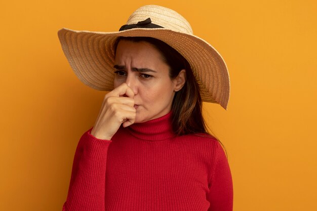 Free photo unpleased pretty woman with beach hat holds nose isolated on orange wall