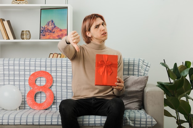 Free photo unpleased showing thumb down handsome guy on happy women day holding present sitting on sofa in living room