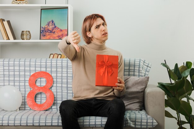 Unpleased showing thumb down handsome guy on happy women day holding present sitting on sofa in living room