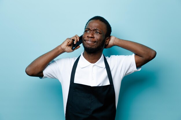 Unpleased speaks on the phone putting hand on head young african american barber in uniform isolated on blue background