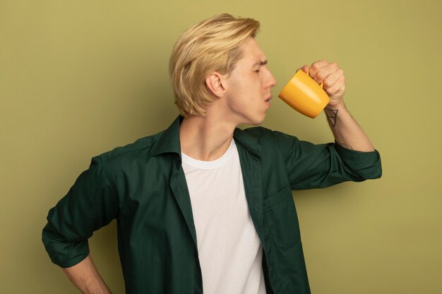 Unpleased with closed eyes young blonde guy wearing green t-shirt drinks tea